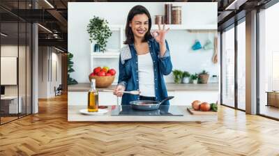 Beautiful young woman looking at camera while cooking healthy food in frying pan in the kitchen at home. Wall mural