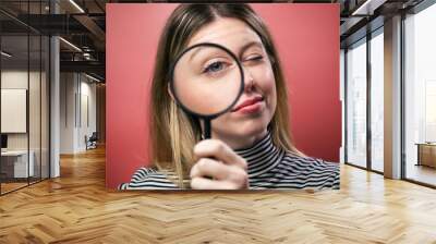 Cheerful young woman looking through magnifying glass at the camera over pink background. Wall mural