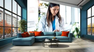 Confident young female doctor reviewing the patient's medical history on her digital tablet while standing in the consultation. Wall mural