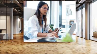 Confident young female doctor using her mobile phone while working on laptop in the consultation. Wall mural