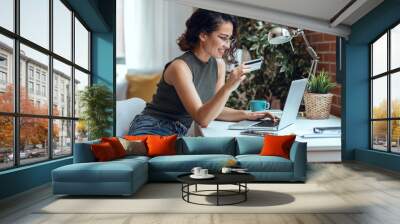 Cute woman holding white credit card for shopping online with computer while sitting in living room at home. Wall mural