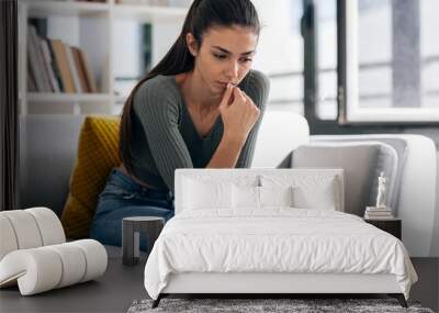 Depressed young woman thinking about her problems while holding mobile phone sitting on the sofa at home. Wall mural