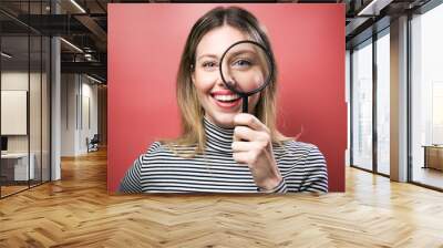 Funny young woman looking through magnifying glass at the camera over pink background. Wall mural