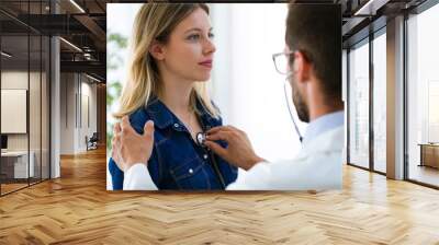 Handsome young male doctor checking beautiful young woman patient heartbeat using stethoscope in medical office. Wall mural