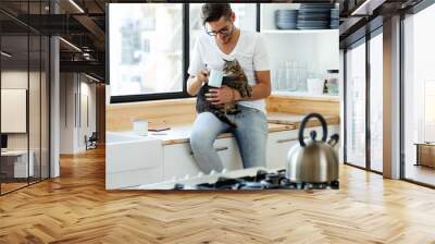 Handsome young man playing with cat in the kitchen. Wall mural