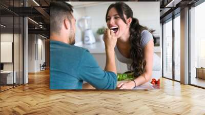 Happy young couple enjoying lunch while handsome man is feeding with yogurt his wife in the kitchen at home. Wall mural