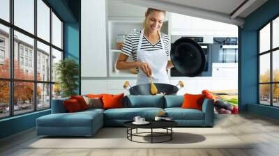 Healthy young woman cooking and mixing food in frying pan in the kitchen at home. Wall mural