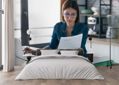 Pretty young business woman working with computer while consulting some invoices and documents in the kitchen at home. Wall mural