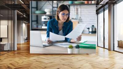 Pretty young business woman working with computer while consulting some invoices and documents in the kitchen at home. Wall mural