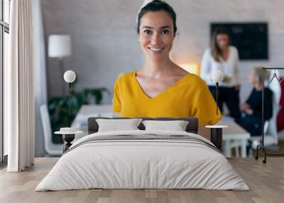 Pretty young businesswoman looking at camera. In the background, her colleagues working in the office. Wall mural