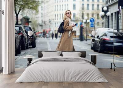 Pretty young woman crossing the street while holding the smartphone and looking sideways. Wall mural