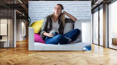 Pretty young woman listening to music with headphones while drinking cup of coffee on sofa at home. Wall mural