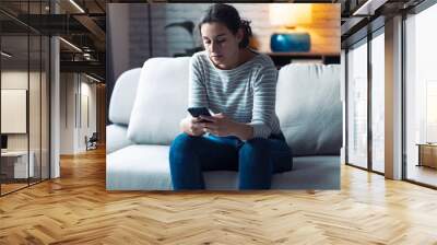 Serious young woman using her mobile phone while sitting on sofa in the living room at home. Wall mural