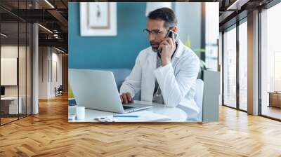 Shot of mature concentrated attractive male doctor working with her computer while talking with smartphone in medical consultation. Wall mural