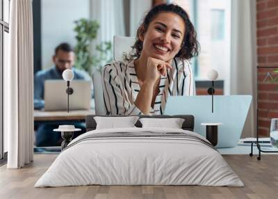 Smiling business woman working with laptop while looking at camera in modern startup office. Wall mural