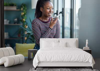 Smiling young african american woman sending audio message with mobile phone while sitting on sofa at home Wall mural