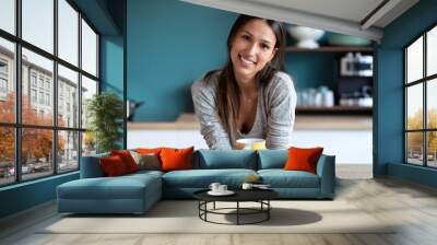 Smiling young woman looking to the laptop web camera while holding a cup of coffee in the kitchen at home. Wall mural