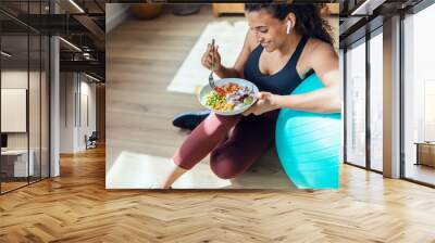 Sporty young woman eating healthy while listening to music sitting on the floor at home. Wall mural