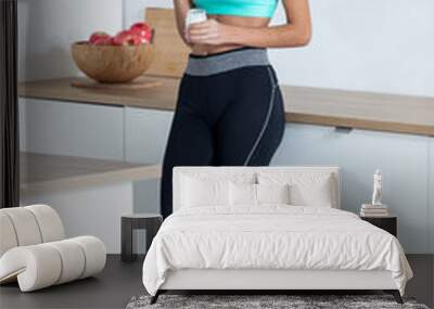 Sporty young woman eating iogurt while standing in the kitchen at home. Wall mural