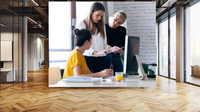 Three modern businesswomen talking and reviewing the latest work done on the digital tablet in a joint workspace. Wall mural