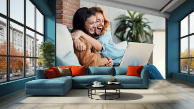 Two happy beautiful women doing a video call with laptop while hugging each other sitting on the couch at home. Wall mural