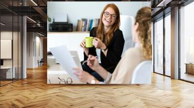 Two pretty young business woman relaxing one moment while drinking coffee in the office. Wall mural