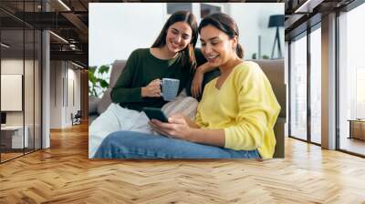 Two smiling young women talking while watching smartphone sitting on couch in the living room at home. Wall mural