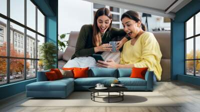 Two smiling young women talking while watching smartphone sitting on couch in the living room at home. Wall mural