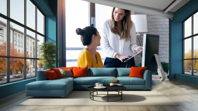 Two young businesswomen talking and reviewing they last work in the digital tablet in the office. Wall mural