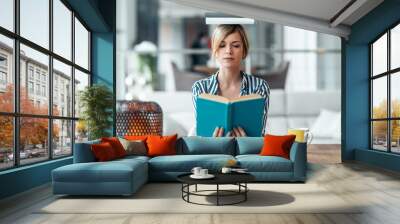 Young attractive woman at the desk with books on her head while reading a book in living room at home. Wall mural