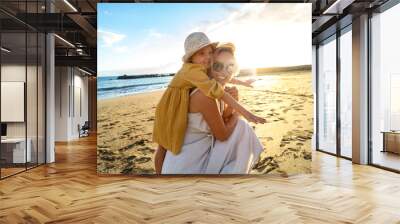 Happy mother and her little daughter having fun together on the sandy beach during sunset. Lovely kid embracing and playing with her mom during summer vacation. Real people emotion. Wall mural