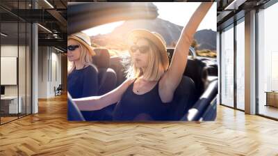 Two happy blonde caucasian girls wearing sunglasses and summer hats driving cabrio car during vacation road trip on sunny day. Freedom. Happiness. Dreams. Tourist. Wanderlust. Wall mural