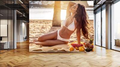 Young woman with beautiful fit tan body having picnic with fresh fruits at the empty tropical beach. Girl with long dark hair and natural look wearing white fashionable bikini. Wall mural