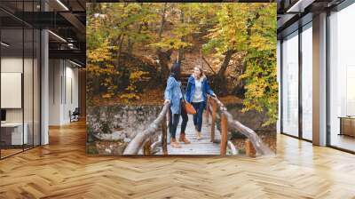 Two young girl friends walking on a wooden bridge in the autumn forest, talking and laughing. Wall mural