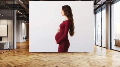 Young beautiful pregnant woman with long curly brunette hair, wearing burgundy dress, holding hands on her belly on white background. Wall mural