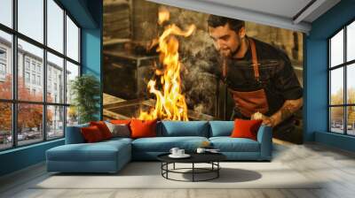 Bearded handsome man in jeans apron and with tattoo on hand cooking. Chef smiling, holding oil in hand and looking at meat on grill. Tasty beef steak flaming with fire and smoke. Wall mural