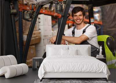 Good looking loader sitting in forklift, posing and smiling. Professional worker wearing uniform and white t shirt. Background of warehouse with many boxes and goods. Wall mural