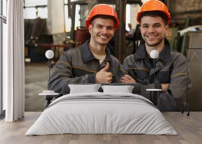 Portrait of two happy workers of metal stock in orange helmets and gray uniform standing together, posing and looking at camera. Man showing sight thumb up and his colleague standing with arms folded. Wall mural