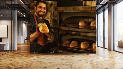 Smiling baker showing fresh baked bread and holding it in his hand. Beautiful and handsome man standing near special convection oven in bakery. Concept of baker's shop. Wall mural