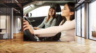 Two beautiful and pretty girls driving in car of their dream. Happy female driver holding hands on steering wheel. Girls smiling because of successful purchase of automobile in car dealership. Wall mural