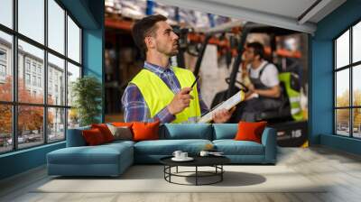 Two workers of warehouse concentrated on their job. Professional manager in reflective waistcoat checking and counting boxes and goods. Forklift driver operating vehicle. Wall mural