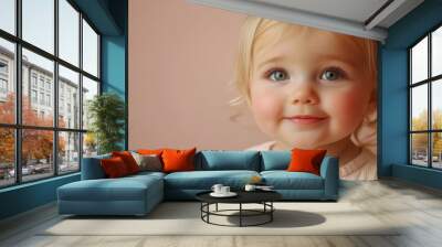 A cute little blond baby girl smiles warmly against a solid beige background, showcasing her bright blue eyes and playful spirit during a joyful moment at home Wall mural