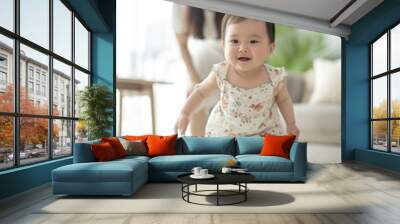 A smiling asian baby beams with pride taking first steps while a supportive mother watches from behind in a bright, cozy living room.. Wall mural