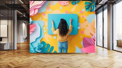 A woman is seen from behind as she interacts with a colorful and whimsical paper art installation featuring playful shapes, creating engaging graphics for social media posts. Social media manager Wall mural