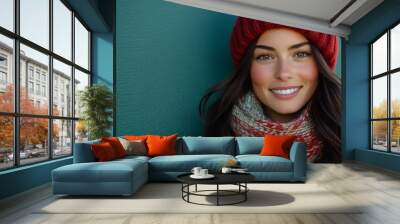 Happy brunette woman in a vibrant red winter hat and scarf against a solid teal background, showcasing her radiant smile and warm demeanor for a cheerful winter vibe Wall mural