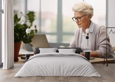 An older woman with glasses sits at a table working on a laptop. There is a potted plant and a cup near the laptop. Wall mural