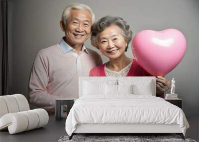 An elderly couple stands side by side in a studio setting holding a pink heart balloon and smiling with warmth and joy celebrating their love and creating cherished memories cherished together Wall mural