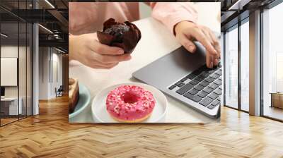 Bad habits. Woman eating muffin while using laptop at white wooden table, closeup Wall mural