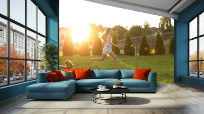 Beautiful girl running with cute Maltipoo dog on green lawn at sunset in backyard Wall mural