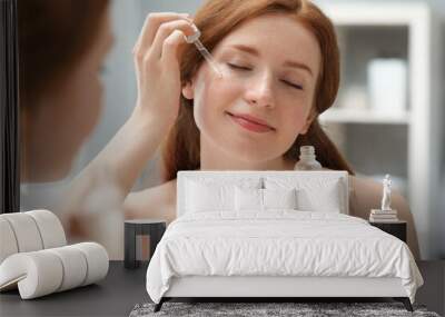 Beautiful woman with freckles applying cosmetic serum onto her face near mirror in bathroom Wall mural